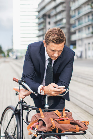 Businessman using smart phone in city while leaning on bicycle stock photo