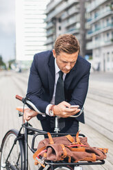 Geschäftsmann benutzt sein Smartphone in der Stadt, während er sich auf ein Fahrrad stützt - MASF05138