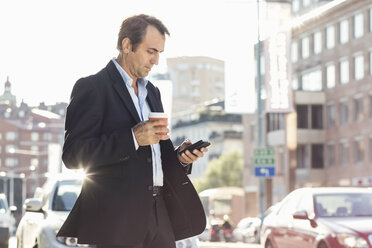 Businessman using mobile phone and holding disposable coffee cup while walking on city street - MASF05146