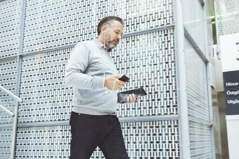 Geschäftsmann mit digitalem Tablet, der beim Spazierengehen im Büro mit seinem Mobiltelefon Musik hört - MASF05158