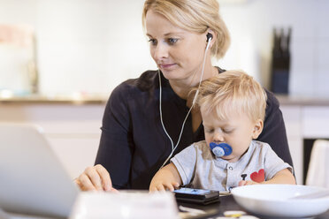 Ein kleiner Junge benutzt ein Smartphone, während die Mutter am Tisch auf dem Laptop Musik hört - MASF05159