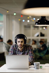 Businessman listening music while working late on laptop in creative office - MASF05162