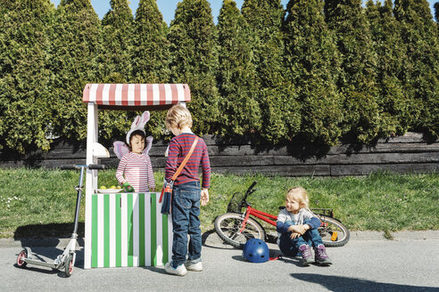 Boy standing at food stall while friend imitating as seller on footpath - MASF05186