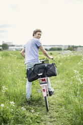 Rückansicht eines jungen Mannes mit Tasche und Fahrrad auf einem Feld stehend - MASF05255