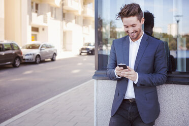 Young businessman using mobile phone against wall by sidewalk - MASF05330