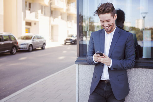 Junger Geschäftsmann, der ein Mobiltelefon an der Wand eines Bürgersteigs benutzt - MASF05330