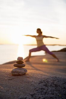 Woman practicing yoga warrior pose on lakeshore with focus on stack of stones - MASF05347