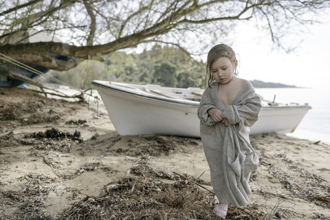 Sad little girl wrapped in oversized cardigan standing on the beach stock photo