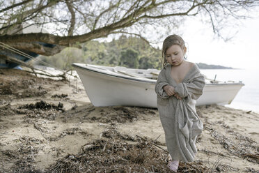 Sad little girl wrapped in oversized cardigan standing on the beach - KMKF00187