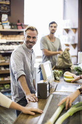 Smiling man standing at supermarket checkout counter with friend in background - MASF05379