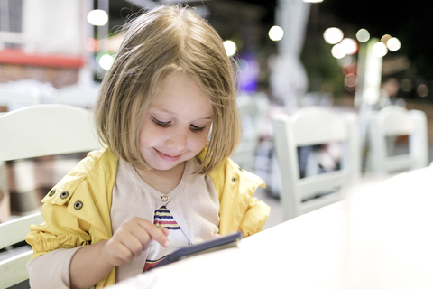 Porträt eines lächelnden kleinen Mädchens, das in einem Restaurant sitzt und mit seinem Smartphone spielt, lizenzfreies Stockfoto