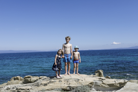 Griechenland, Chalkidiki, drei Kinder strampeln Hand in Hand auf einem Felsen vor dem Meer, lizenzfreies Stockfoto