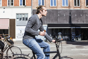 Profilaufnahme eines jungen Mannes, der einen Burger in der Hand hält, während er mit dem Fahrrad durch die Stadt fährt - MASF05430