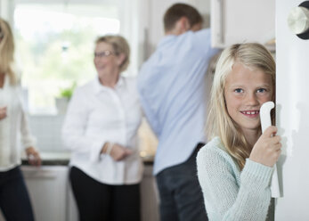 Porträt eines lächelnden Mädchens, das die Türklinke hält, mit Familie im Hintergrund in der Küche - MASF05444