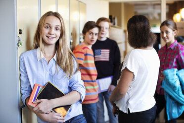 Portrait of school girl standing at locker room with friends in background - MASF05455