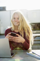 Porträt eines glücklichen Teenagers mit Mobiltelefon und Laptop am Tisch im Freien - MASF05458