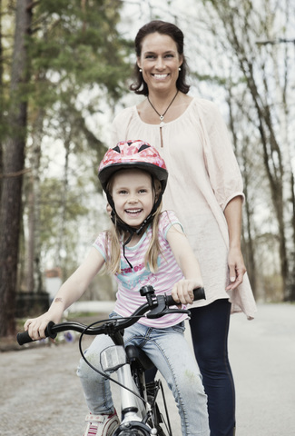 Porträt eines glücklichen Mädchens auf dem Fahrrad, während die Mutter hinter ihr auf der Straße steht, lizenzfreies Stockfoto