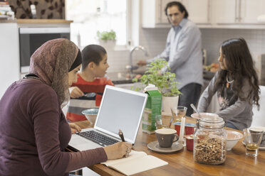 Muslimische Frau arbeitet am Laptop, während ihre Familie in der Küche frühstückt - MASF05534