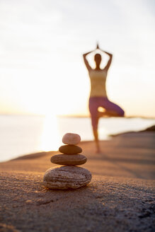 Pile of stones with woman practicing yoga in background at lakeshore - MASF05536