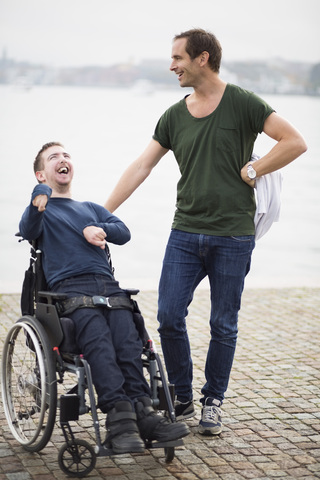 Male caretaker standing with happy disabled man on wheelchair by lake stock photo