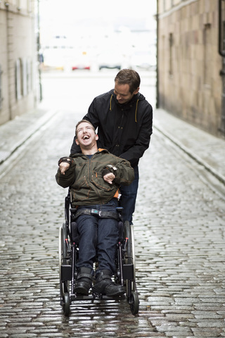 Männlicher Hausmeister mit behindertem Mann im Rollstuhl auf einer Straße in der Stadt, lizenzfreies Stockfoto