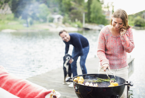 Glückliche Frau, die ein Mobiltelefon benutzt, während sie mit einem Mann und einem Hund im Hintergrund auf einem Pier grillt, lizenzfreies Stockfoto