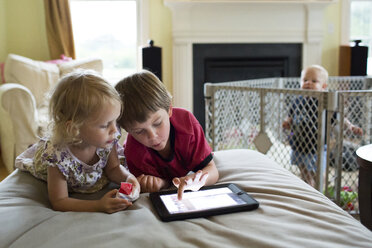 Ein kleiner Junge sieht seine Geschwister an, die einen Tablet-Computer benutzen, während er in seinem Kinderbett steht - CAVF43027