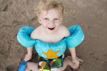 Porträt eines glücklichen Jungen mit Schwimmflügeln, der am Sandstrand sitzt - CAVF43077