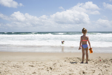 Rear view of girl looking at brother playing in sea against cloudy sky - CAVF43083