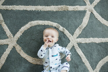Overhead portrait of cheerful baby boy lying on carpet at home - CAVF43091
