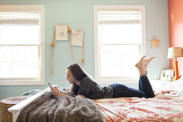 Seitenansicht einer Frau, die ein Mobiltelefon benutzt, während sie zu Hause auf dem Bett liegt - CAVF43093