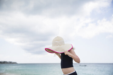 Side view of girl holding hat while standing against sea and cloudy sky at beach - CAVF43160
