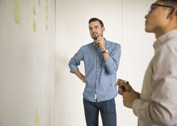 Geschäftsleute, die über Haftnotizen diskutieren, während sie an einer weißen Wand in einem kreativen Büro stehen - CAVF43243