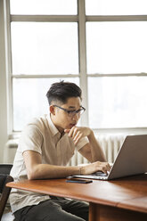 Businessman using laptop while sitting at table against window in brightly lit creative office - CAVF43263