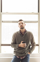 Businessman using mobile phone while leaning on window sill at brightly lit office - CAVF43265