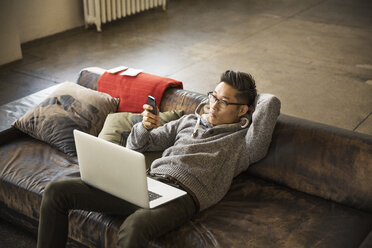 High angle view of businessman with laptop computer using phone while reclining on sofa in creative office - CAVF43289