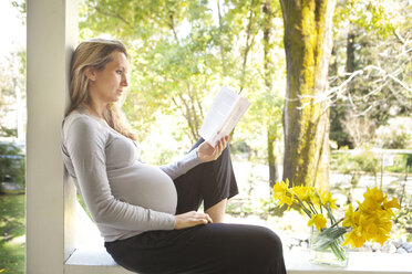 Schwangere Frau liest ein Buch, während sie sich auf der Veranda an einem sonnigen Tag ausruht - CAVF43319