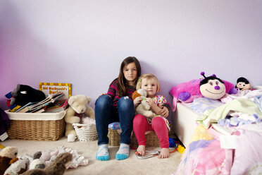 Porträt von Schwestern, die inmitten von Spielzeug an einer Wand im Schlafzimmer sitzen - CAVF43356