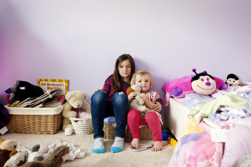 Porträt von Schwestern, die inmitten von Spielzeug an einer Wand im Schlafzimmer sitzen - CAVF43356