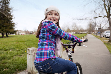 Happy girl cycling on street by grassy field against sky - CAVF43357