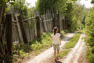 Rückansicht eines Mädchens, das an einem sonnigen Tag auf einem unbefestigten Weg durch einen Holzzaun läuft - CAVF43455