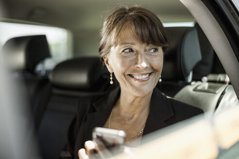 Glückliche Geschäftsfrau mit Mobiltelefon in der Hand, die durch ein Taxifenster hinausschaut - MASF05607