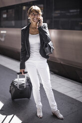 Happy businesswoman with suitcase using mobile phone on railroad station platform - MASF05610