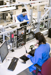 Female technician working on machine part at desk with colleague in background - MASF05660