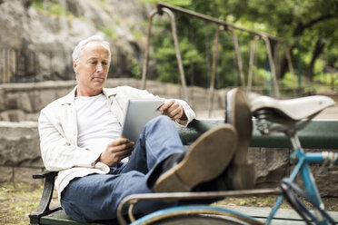 Businessman with feet up on bicycle using digital tablet on park bench - MASF05727