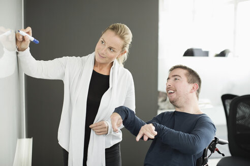 Geschäftsfrau schreibt auf Whiteboard bei der Arbeit mit behindertem Geschäftsmann im Büro - MASF05859