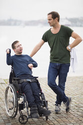 Behinderter Mann im Rollstuhl mit männlichem Pfleger am See - MASF05861