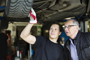 Junger Mechaniker, der mit einem Kunden unter dem Auto in der Werkstatt über Reifen diskutiert - MASF05931