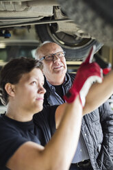 Junger Mechaniker, der mit einem Kunden unter dem Auto in der Werkstatt über Reifen diskutiert - MASF05932