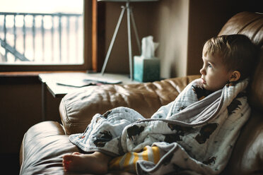 Thoughtful boy wrapped in blanket relaxing on sofa at home - CAVF43465